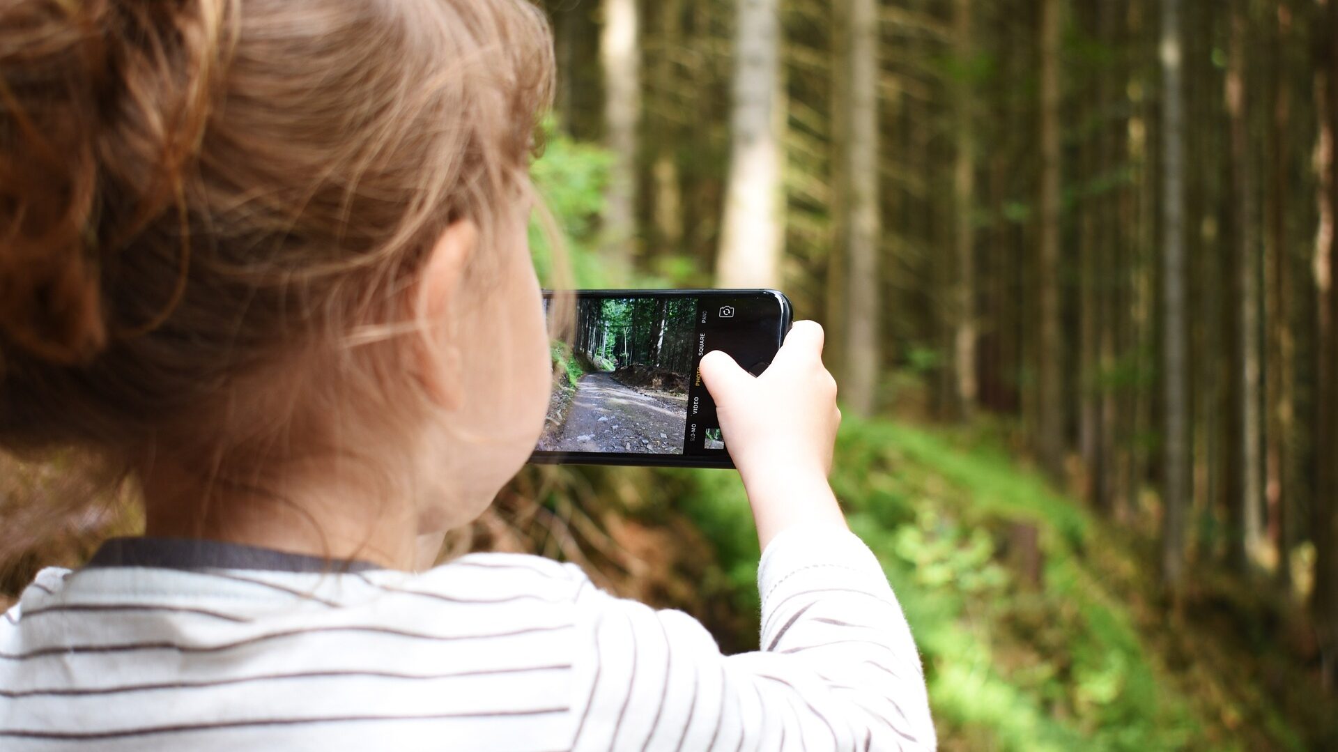 kleines Kind in Rückansicht, das mit einem Handy einen Waldweg fotografiert