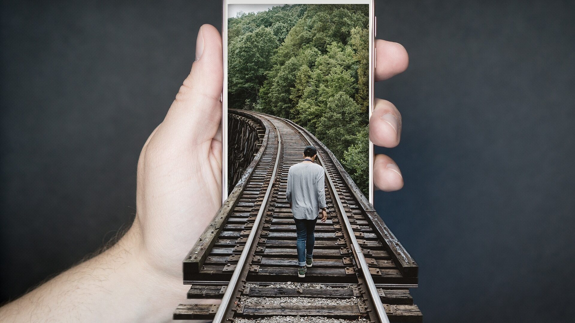 Symbolbild Outdoor Handy - Collage eines Mannes, der über ein Gleis in ein von einer Hand gehaltenen Zugstrecke in das Handy hineinläuft